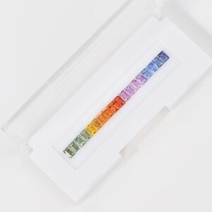 May include: A collection of multi-coloured gemstones arranged in a line within a white case. The gemstones transition from green to violet. The case is open, showing the gemstones.