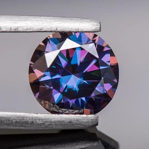 May include: A round, faceted gemstone with a purple and blue colour gradient. The gemstone is held in a pair of tweezers.