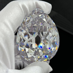 Könnte beinhalten: Ein großer, birnenförmiger, klarer facettierter Edelstein mit Brillantschliff, der das Licht in verschiedene Richtungen reflektiert und ein Farbspektrum erzeugt. Der Stein wird von einer Hand in einem weißen Stoffhandschuh vor dunklem Hintergrund gehalten.