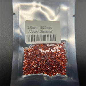 Puede incluir: Un primer plano de una bolsa de 1000 cristales de circonita roja AAA de 2,0 mm. La bolsa está etiquetada con el tamaño y la cantidad de los cristales.