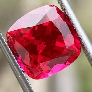 May include: A vibrant red gemstone with a cushion cut. The gemstone is faceted and has a high polish.
