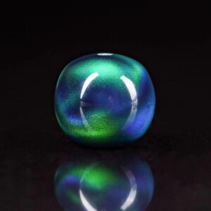 May include: A spherical glass ornament, displaying a mix of blue and green colours. The glass has an iridescent quality, reflecting light. The object is positioned on a dark surface, with its reflection clearly visible.