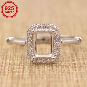 Könnte beinhalten: Ein Ring aus 925er Sterlingsilber mit einer rechteckigen Fassung für einen Mittelstein. Der rechteckige Rahmen ist von einem Kranz aus kleinen, klaren, rund geschliffenen Steinen umgeben. Die Ringschiene ist glatt und poliert mit vier Krappen.