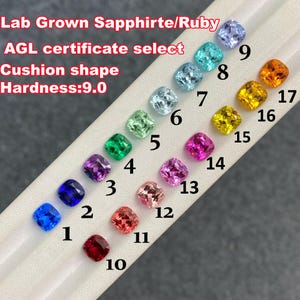 May include: A selection of lab-grown sapphires and rubies in various colours, such as blue, green, pink, and yellow. The cushion-shaped gemstones are displayed on a white surface. The image text states "Lab Grown Sapphirte/Ruby" and "AGL certificate select".