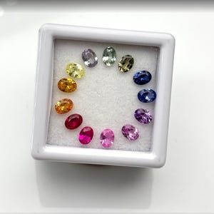 May include: A selection of oval-shaped gemstones in a spectrum of colours, including yellow, orange, red, pink, purple, blue, and green, arranged in a circular pattern within a white display case. The gemstones are displayed against a white background.