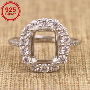 May include: A 925 silver ring featuring a rectangular center setting surrounded by a halo of small, clear, round-cut stones. The band is thin and also accented with small, clear, round-cut stones along the sides. The text 925 Silver is visible.