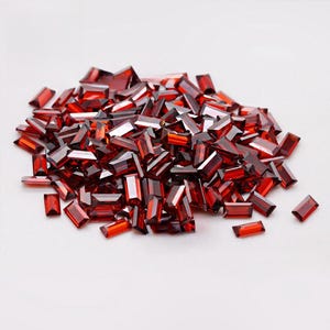 May include: A large collection of small, deep red, rectangular baguette-cut gemstones. Each individual stone features a faceted surface that reflects light, creating a vibrant and uniform appearance across the entire pile of loose, polished gems.