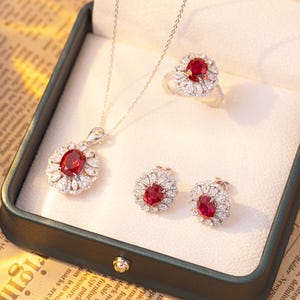 May include: A jewelry set consisting of a necklace, a ring, and a pair of stud earrings. Each piece features a central oval-cut red gemstone surrounded by a halo of small, clear, marquise and round-cut stones set in a metallic frame.