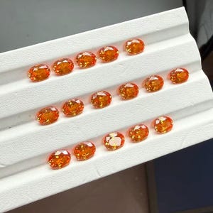 May include: A collection of oval-cut, orange-colored gemstones arranged on a white surface. The gemstones have a brilliant, faceted appearance, reflecting light. The arrangement suggests a display of precious stones, possibly for jewelry making or collecting.