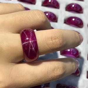 Puede incluir: Un anillo de piedra preciosa rojo oscuro con un patrón en forma de estrella. El anillo es de un color rico y brillante y parece estar engastado en una banda lisa. Otros anillos similares son visibles en el fondo, sugiriendo una colección de joyas.