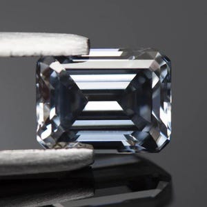 Puede incluir: Una gema de talla esmeralda azul claro sostenida por unas pinzas. La gema tiene muchas facetas que reflejan la luz. La gema rectangular tiene esquinas cortadas y un patrón geométrico.