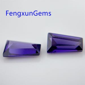 Puede incluir: Dos cabujones de piedra preciosa facetados de color púrpura, cortados en forma rectangular con un extremo puntiagudo. Las piedras preciosas están sobre una superficie blanca.