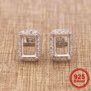 May include: A pair of rectangular stud earrings crafted from 925 silver, featuring a central open setting surrounded by a halo of small, clear, round-cut stones. Each earring has a post back and a polished metallic finish with clear detailing.
