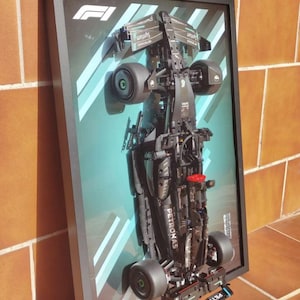 Puede incluir: Modelo de coche de carreras de Fórmula 1 negro enmarcado. El modelo es negro con la marca Petronas y detalles rojos. El fondo es un degradado turquesa con rayas diagonales blancas. El marco es negro.