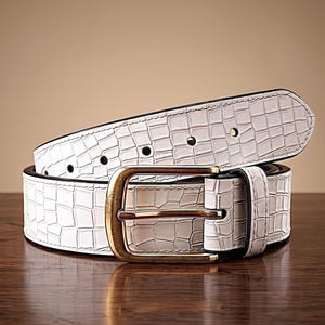 May include: White belt with a textured, crocodile-like pattern. The belt features a gold-colored buckle and a black edge. The belt is displayed on a wooden surface.