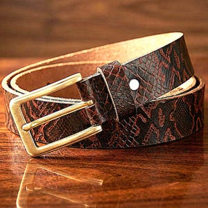 Puede incluir: Un cinturón de cuero marrón enrollado con un estampado de piel de serpiente en marrón oscuro y rojo. El cinturón tiene una hebilla rectangular dorada y un solo remache blanco. El cinturón se muestra sobre una superficie de madera.