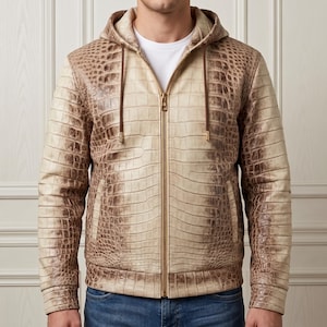 Op de afbeelding: Een beige en bruine jas met capuchon en een textuur van krokodillenleer. De jas heeft een goudkleurige ritssluiting over de gehele lengte, lange mouwen en twee zijzakken. Donkerbruine trekkoorden met goudkleurige uiteinden hangen aan de capuchon.