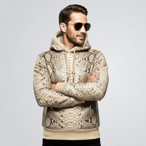 Pode incluir: Um moletom bege com estampa de pele de cobra, capuz com cordão e punhos canelados. O moletom tem uma paleta de cores neutras com tons de bege e marrom. O texto "huzi clothes" é visível na bainha inferior.