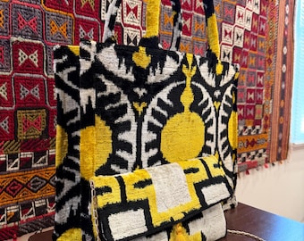 May include: A tote bag and a clutch with a yellow, black, and white patterned design. The tote bag has black and yellow handles. The clutch has a gold chain strap and a yellow tassel. The bags are on a wooden surface.