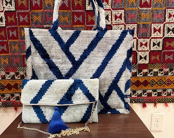 May include: A tote bag and a clutch bag with a geometric pattern in navy blue on a gray background. The clutch has a gold chain strap and a blue tassel. The bags are on a brown table with a patterned textile in the background.
