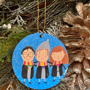May include: A circular Christmas tree decoration with a blue background and a design of three cartoon figures in black robes and scarves. The ornament has a gold hanging string. The figures are holding hands.