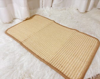 May include: A light brown woven cat scratching mat with a brown stitched border. The mat is on a white fluffy surface.