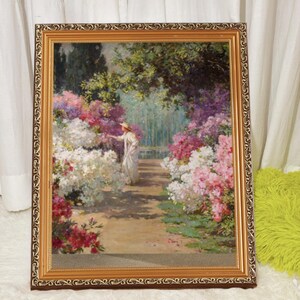 May include: A framed painting depicting a woman in a white dress and hat standing in a garden with a variety of flowers, including white, pink, and purple blossoms.