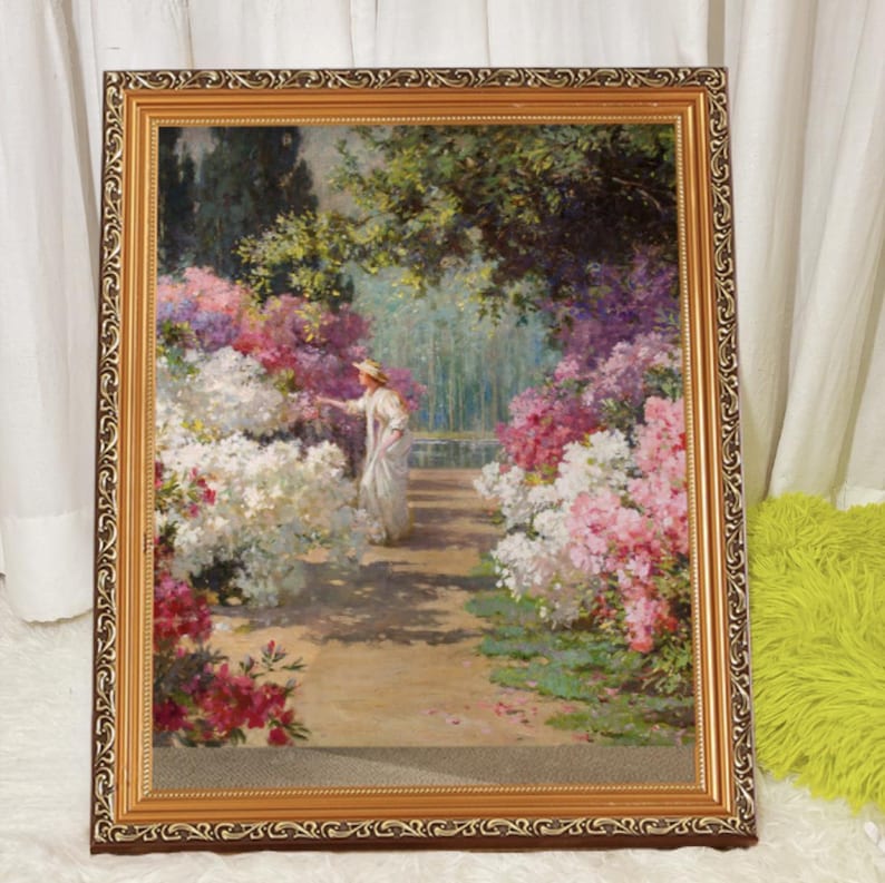 May include: A framed painting depicting a woman in a white dress and hat standing in a garden with a variety of flowers, including white, pink, and purple blossoms.