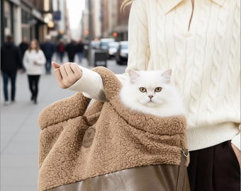 May include: A fluffy white cat peers out of a brown and tan pet carrier. The carrier has a soft, sherpa-like lining and a brown leather-look bottom. The cat carrier is being held outdoors on a city street.