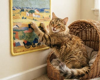 Puede incluir: Un gato atigrado marrón alcanza una pequeña tapicería rectangular que representa un paisaje rural con un borde amarillo. El gato está sentado en una cama para mascotas de mimbre. La tapicería está montada en una pared beige.