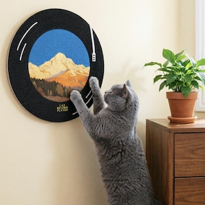 May include: A round black cat scratcher designed to look like a record player, with a mountain landscape in the center. The words "CAT RECORD PLAYER" are printed on the bottom left. A gray cat is reaching up to scratch the surface.