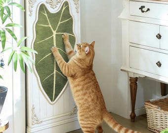 May include: A vertical cat scratching post featuring a white frame with ornate decorative molding and a central green leaf-shaped sisal panel with light-colored veins. The scratching surface is shaped like a large tropical leaf.