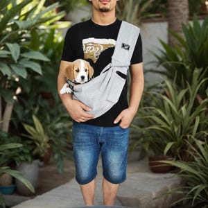 May include: A gray pet carrier sling holds a small beagle puppy. The sling is worn across the body, with the puppy's head visible. The person is wearing a black t-shirt and blue denim shorts.