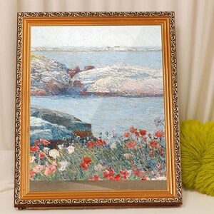 May include: A framed painting depicting a seascape with a rocky shoreline and a field of red poppies in the foreground. The water is a light blue color and the sky is a pale blue. The painting is framed in a gold frame.