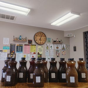 May include: A row of 14 glass bottles filled with a dark brown liquid, sitting on a wooden table. The bottles have white labels with black text. The bottles are in a room with a clock on the wall and several posters.