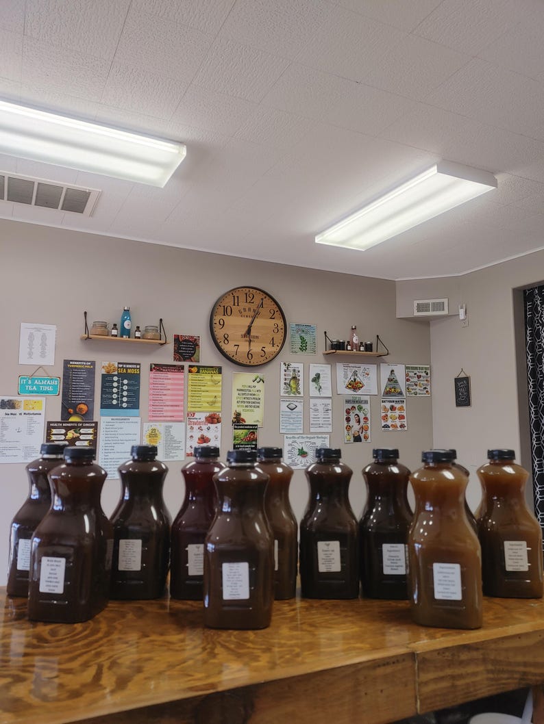 May include: A row of 14 glass bottles filled with a dark brown liquid, sitting on a wooden table. The bottles have white labels with black text. The bottles are in a room with a clock on the wall and several posters.