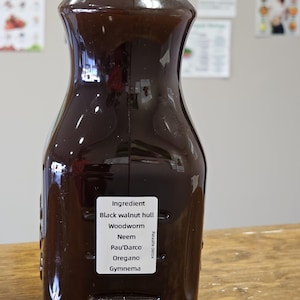 May include: A dark brown glass bottle with a black cap, labeled "Ingredient: Black walnut hull, Woodworm, Neem, Pau'dArco, Oregano, Gymnema.  Parasite detox."
