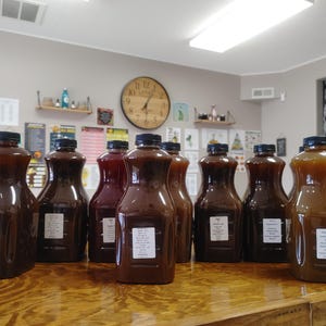 May include: A row of 10 glass bottles filled with various dark liquids, likely homemade syrups or sauces. The bottles are lined up on a wooden table in a room with a clock on the wall.