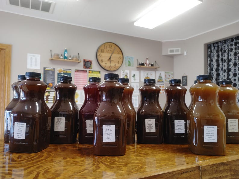 May include: A row of 10 glass bottles filled with various dark liquids, likely homemade syrups or sauces. The bottles are lined up on a wooden table in a room with a clock on the wall.