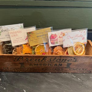 Peut inclure: Une caisse en bois remplie de fruits secs et d'épices, étiquetée "Breakstone's American Process Cheese". La caisse contient quatre petits sachets de pot-pourri avec des étiquettes qui indiquent "Breath Fresh", "Pumpkin Spice", "Farmhouse Simmer" et "Apple Crisp Medley".