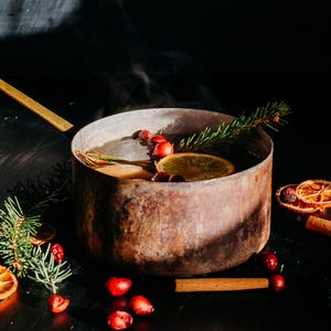 Pode incluir: Uma panela de metal marrom rústica com uma alça dourada cheia de sidra quente fumegante. A panela é cercada por ramos de abeto, paus de canela, fatias de laranja secas e bagas vermelhas.