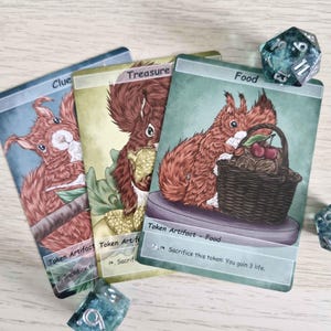 Może przedstawiać: Trzy karty do gry fantasy z ilustracjami wiewiórek. Karty są oznaczone jako "Clue", "Treasure" i "Food". Widoczna jest kostka d20. Karta "Food" przedstawia wiewiórkę z koszem wiśni.