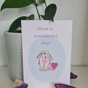 Può includere: Un coniglio bianco e grigio con cuori rosa sul petto tiene un mazzo di cuori rosa. La carta dice "Have a wonderful day!"