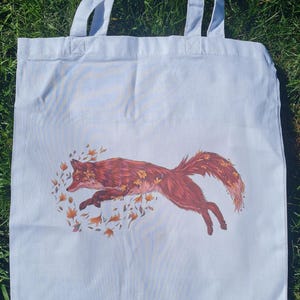 Op de afbeelding: Een witte tote bag met een rode vos die door een cirkel van oranje bladeren springt. De vos is gestileerd afgebeeld, bijna als een cartoon.