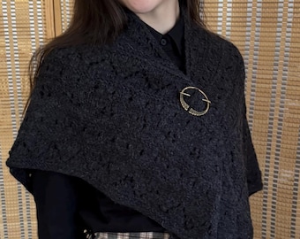 May include: A dark gray knitted shawl with a decorative gold-colored brooch. The shawl is draped over a black shirt and plaid skirt. The brooch is circular with an intricate design.