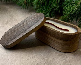 Pode incluir: Uma vela de madeira em um recipiente oval com uma tampa correspondente. A vela tem cera branca e um pavio vermelho. O recipiente e a tampa são feitos de madeira marrom clara. Ramos de pinheiro verdes ao fundo.