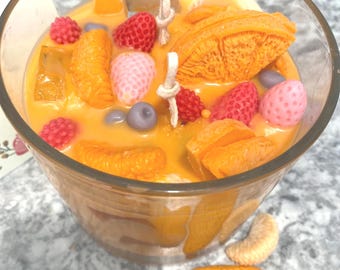 Könnte beinhalten: Ein klares Glasgefäß gefüllt mit einer gelben Wachskerze. Die Kerze ist mit orangefarbenen, rosa und violetten, fruchtigen Wachsstückchen verziert. Das Gefäß steht auf einer grauen Oberfläche.