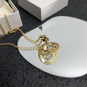 Puede incluir: Un collar con colgante dorado con un diseño en forma de planeta, con un orbe de cristal transparente y una cruz decorativa. El collar se muestra sobre una superficie reflectante, con una caja blanca en el fondo.