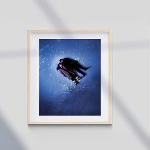 Puede incluir: Impresión artística enmarcada que muestra a dos personas tumbadas sobre una superficie azul, posiblemente hielo, vista desde arriba. Llevan abrigos oscuros y botas. La obra está enmarcada en un marco de madera clara.