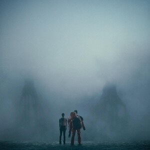 May include: Three figures in a misty environment. One person is in an orange jumpsuit and backpack, the others in dark clothing. The background shows large, shadowy forms, all shrouded in a dense fog.